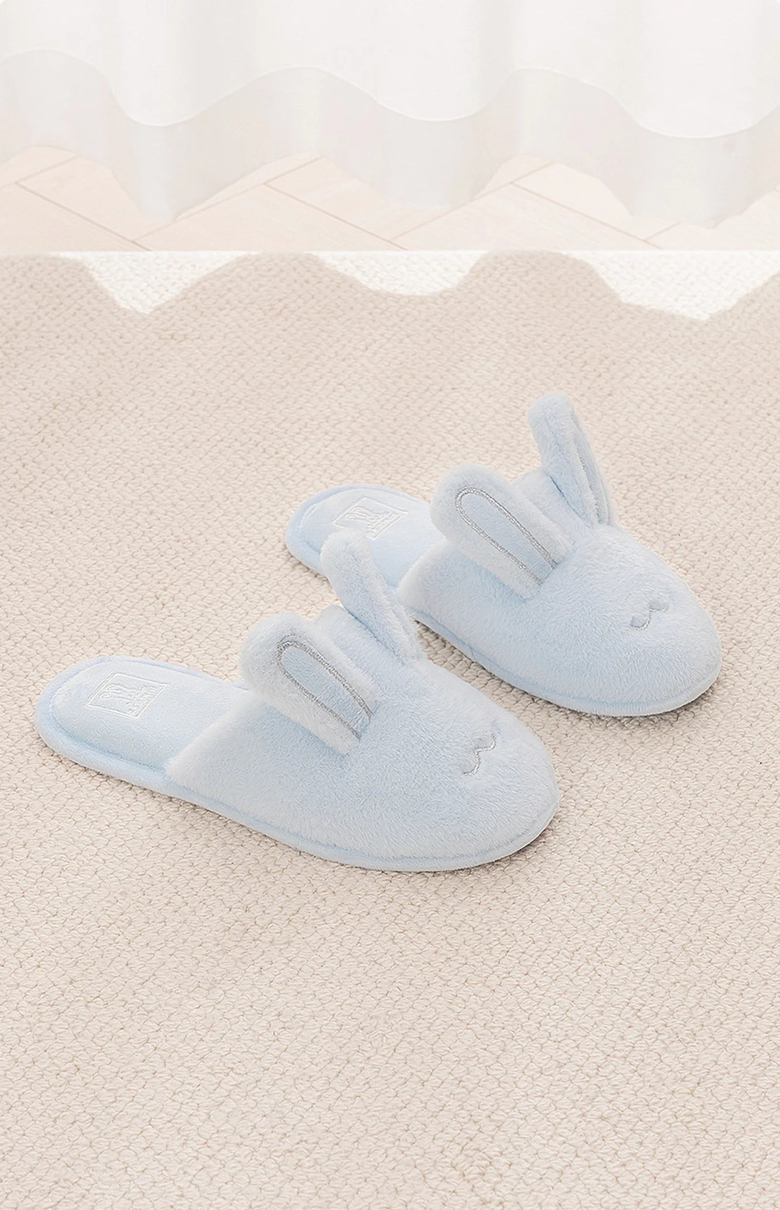 Pair of blue plush bunny slippers on beige rug - RoomDen
