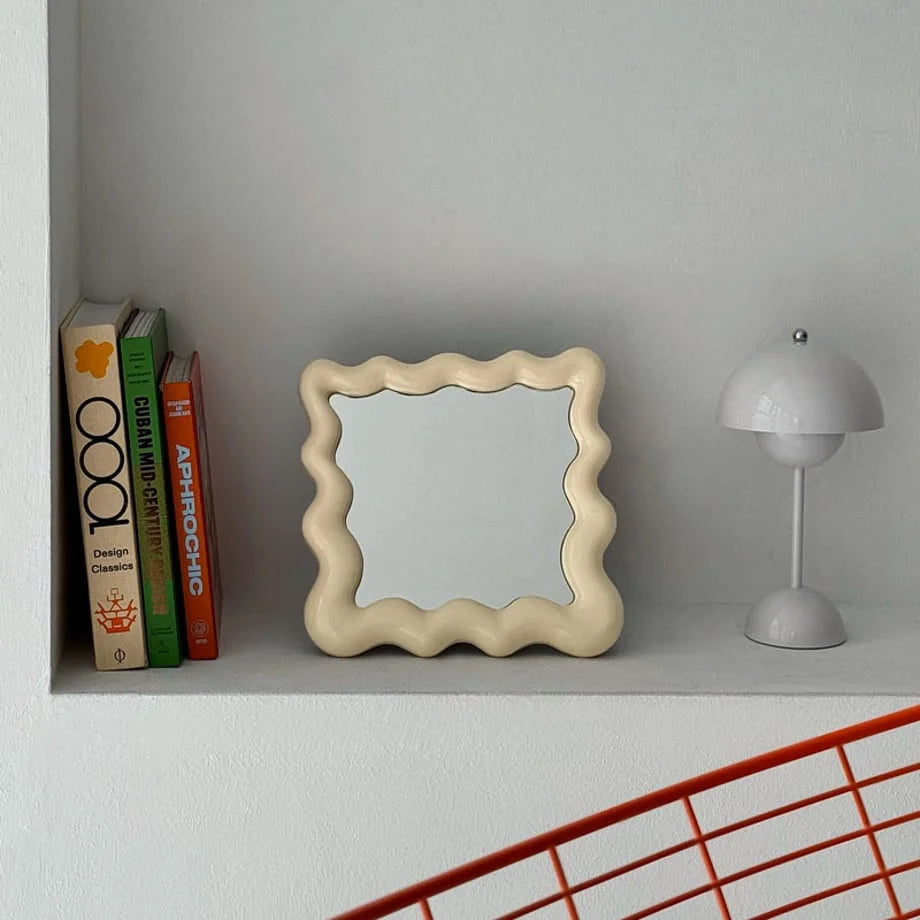 Cream wavy frame desktop mirror resting on a white shelf next to books and a small lamp - RoomDen