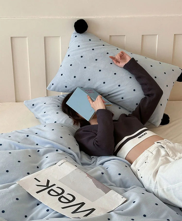 A person relaxing in bed with the Baby Blue Polka Dot Pom Pom Velvet Bedding Set, reading a book. - RoomDen