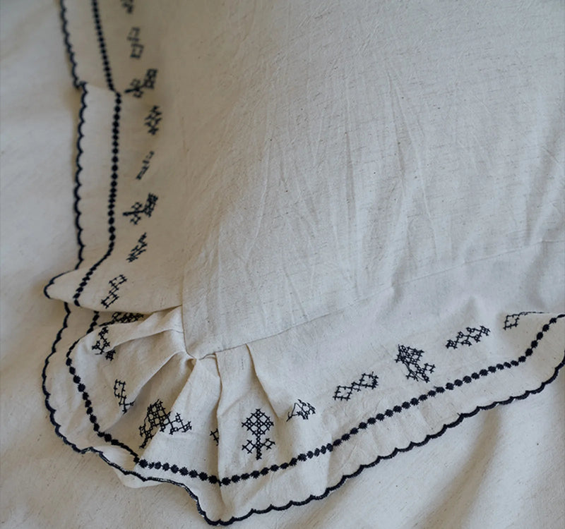 Detailed close-up of the black embroidery and scalloped ruffle on the linen pillowcase corner. — RoomDen