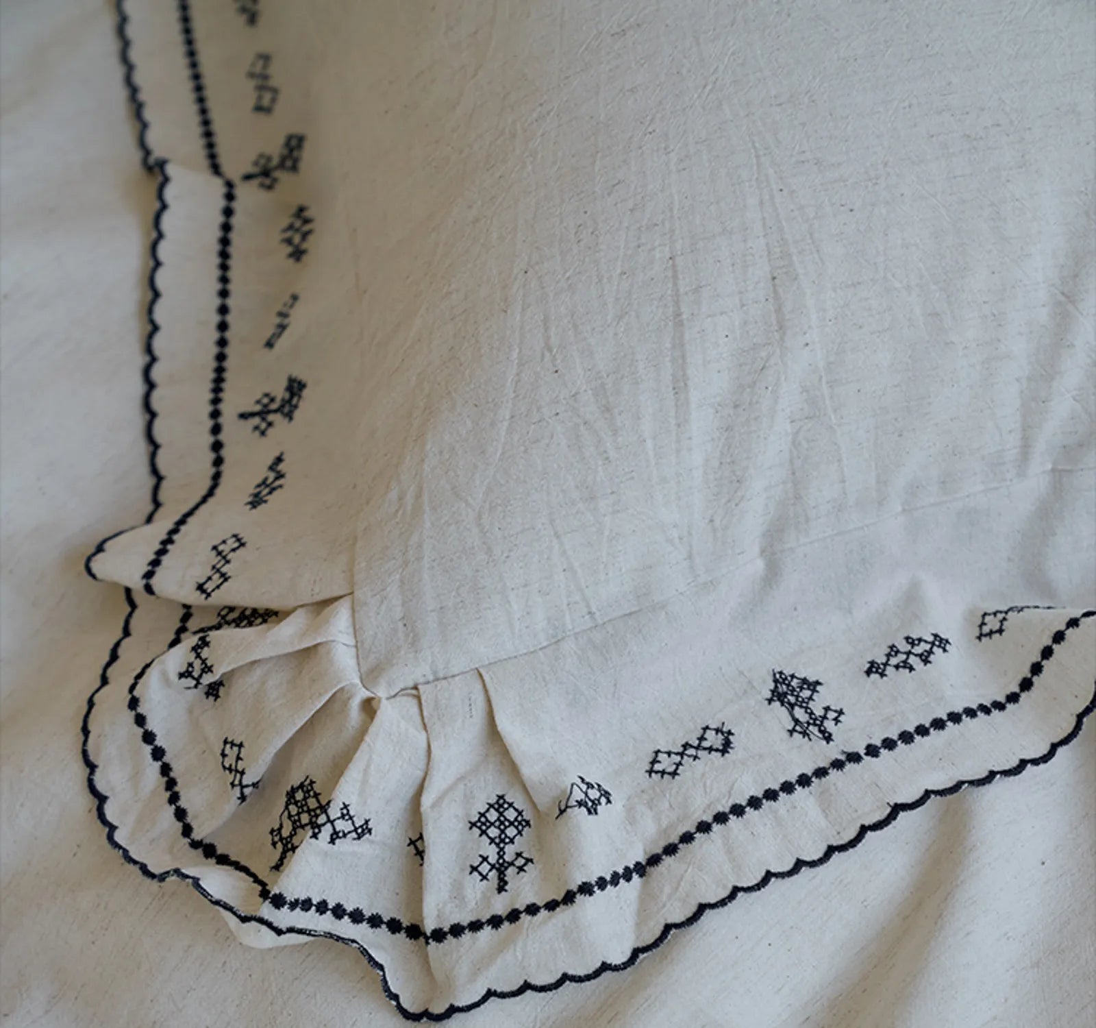 Detailed close-up of the corner of a pillowcase, highlighting the black embroidery and scalloped ruffle design on the linen fabric - RoomDen.