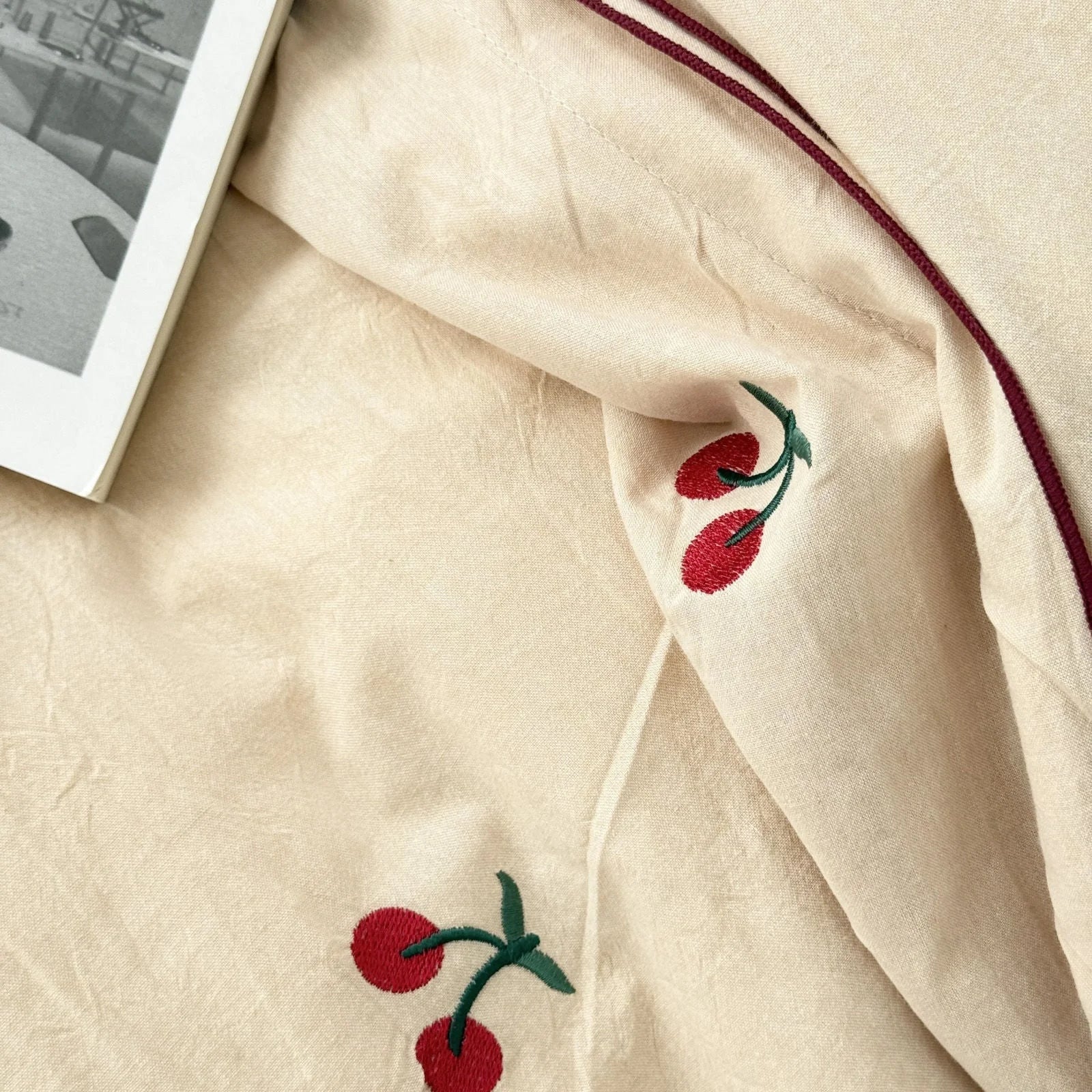Close-up detail of the red and green embroidered cherry on the textured creamy beige washed cotton fabric.
