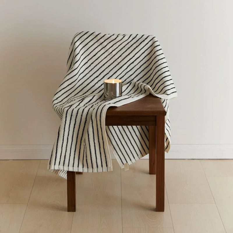 A striped cotton towel over a wooden stool beside a single lit candle in warm evening light, a calm coastal bedroom corner. — RoomDen