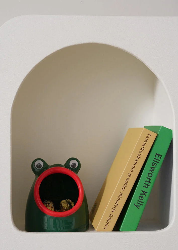A green googly eye frog ceramic organizer tucked inside a white arched shelf beside two books. - RoomDen