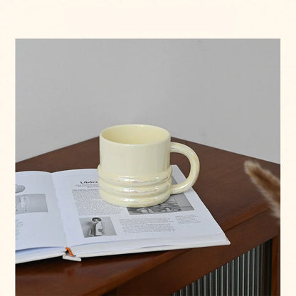 A chunky ribbed ceramic mug in iridescent white resting on an open book on a wooden table - RoomDen.
