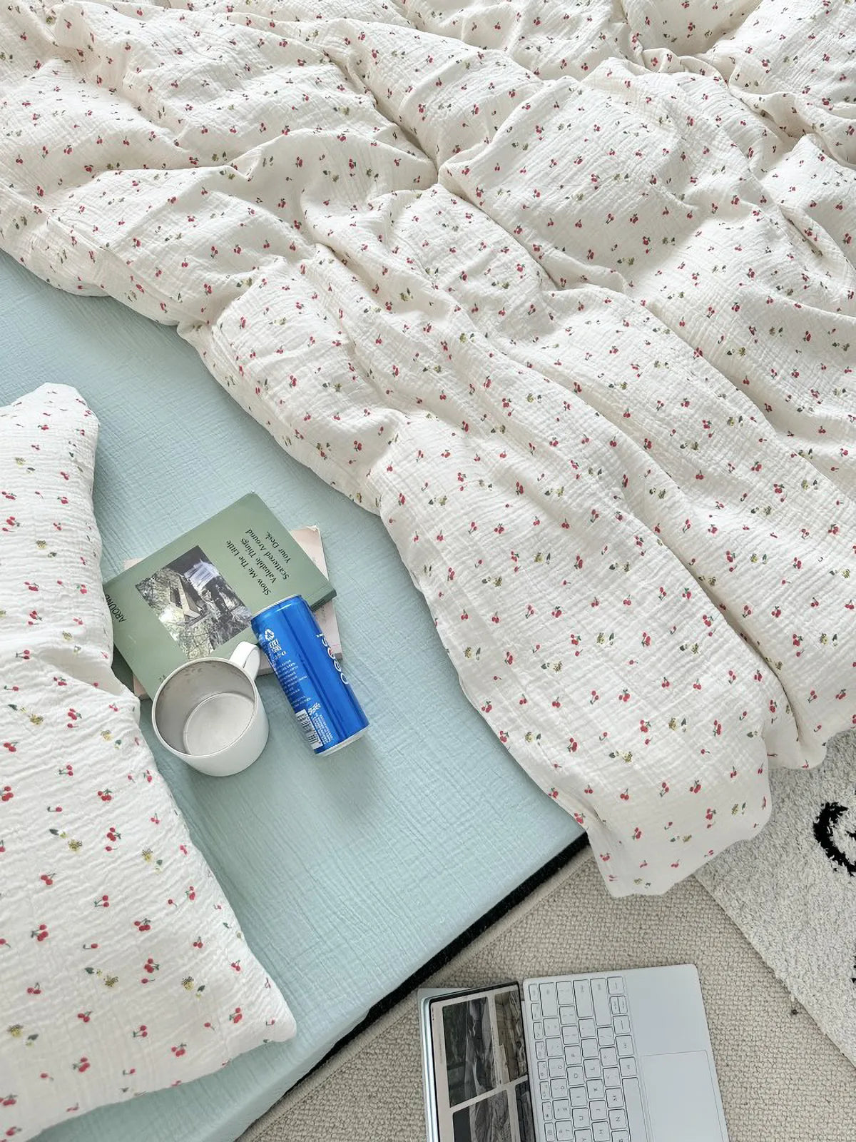 Top-down view of the cherry print bedding set on a bed with a light blue fitted sheet, styled with a laptop, mug, and book. - RoomDen