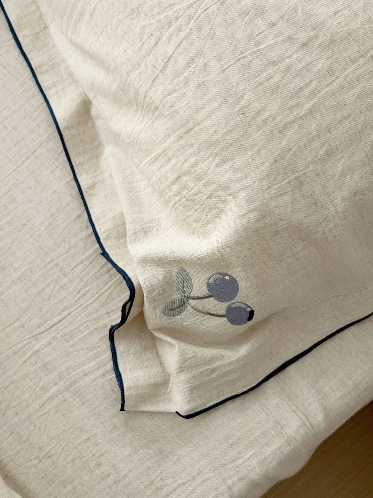 Macro detail shot of the delicate blueberry embroidery and dark piping on the corner of a cream-colored linen pillowcase. - RoomDen