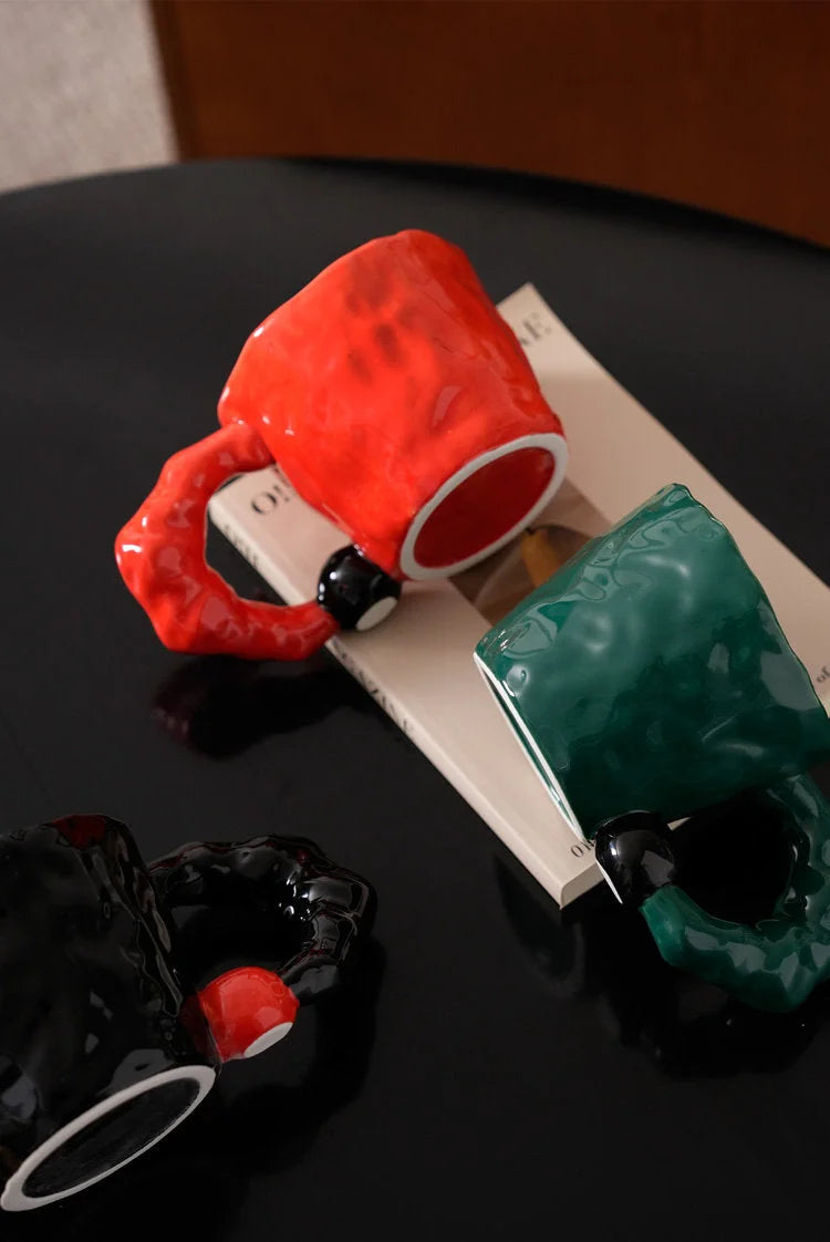 Angled view showing the red, green, and black irregular ceramic mugs on a dark surface - RoomDen.