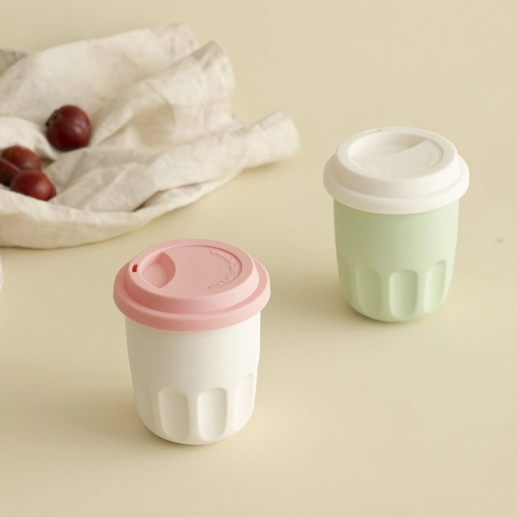 A cream white and a mint green frosted ceramic coffee cup with lids, styled on a table with a linen cloth. - RoomDen