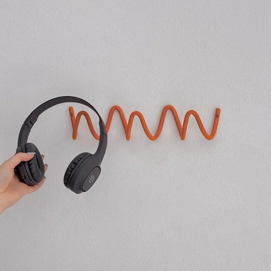 Hand holding headphones next to the orange Wavy Coil Metal Wall Hook. - RoomDen