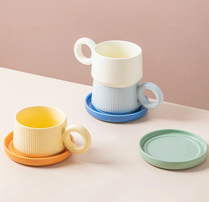 A collection of three ribbed ceramic donut handle mugs in Cream, Blue, and Yellow, stacked and arranged on a table. - RoomDen