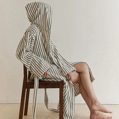 Side profile of a woman sitting on a wooden chair wearing a plush cream and black striped hooded robe.