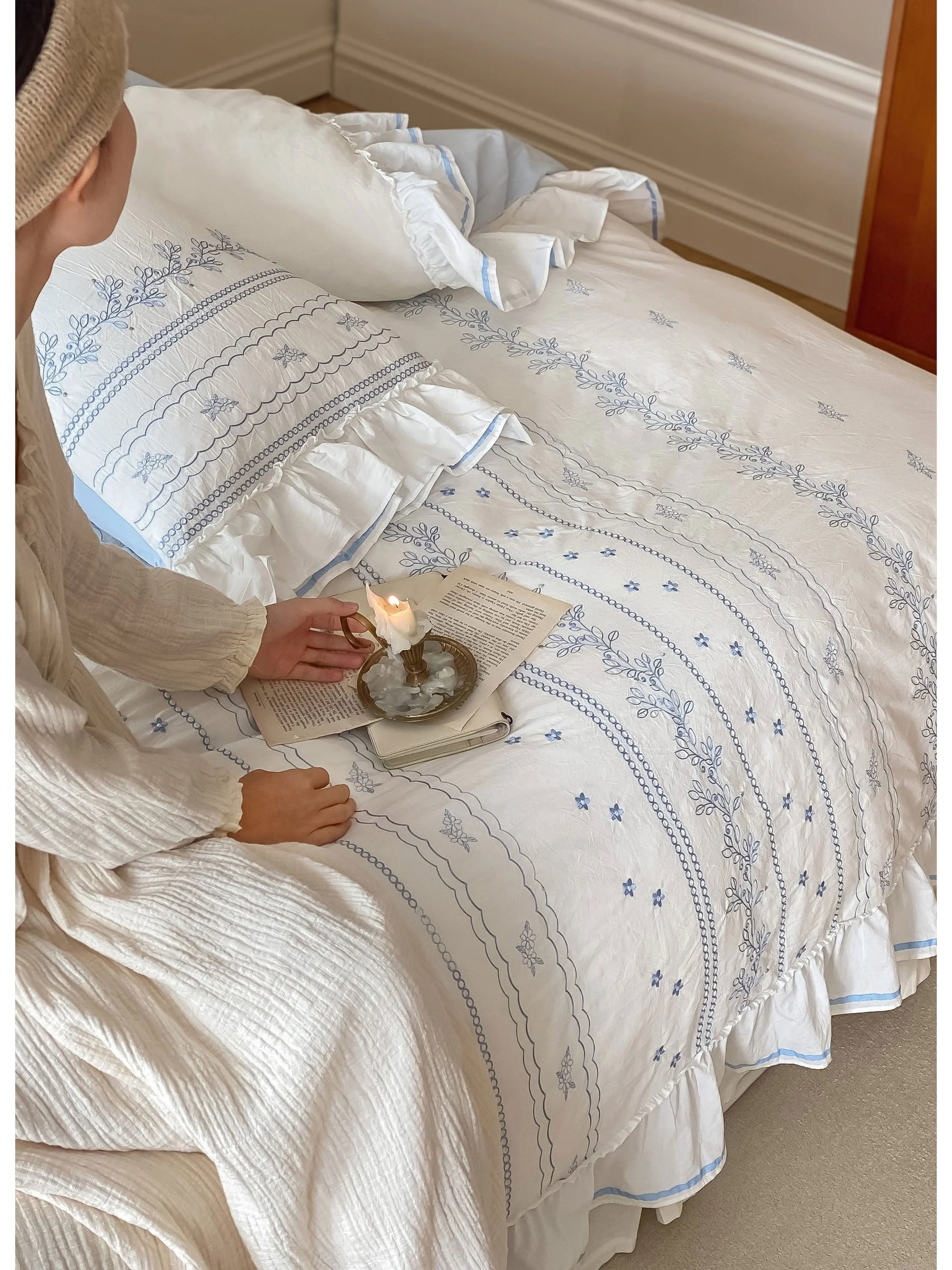 A person holding a candle while sitting on the bed with the Ruffled Floral Vine Embroidered Cotton Bedding Set.