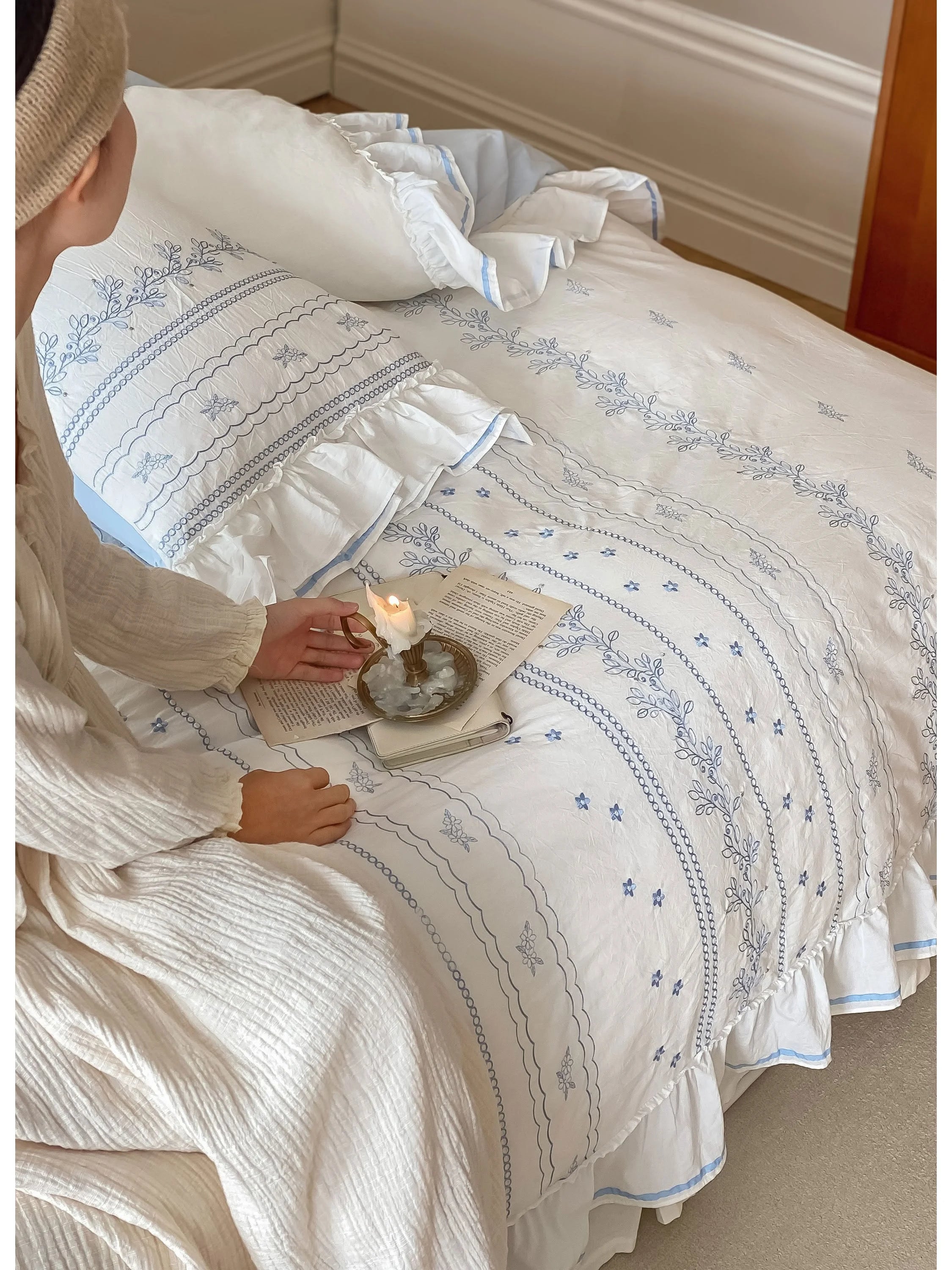 A person holding a candle while sitting on the bed with the Ruffled Floral Vine Embroidered Cotton Bedding Set.