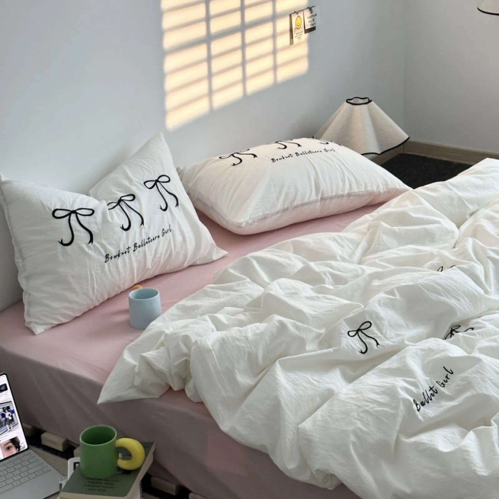The white embroidered bowknot bedding set styled on a bed with a pink fitted sheet, bathed in warm sunlight.