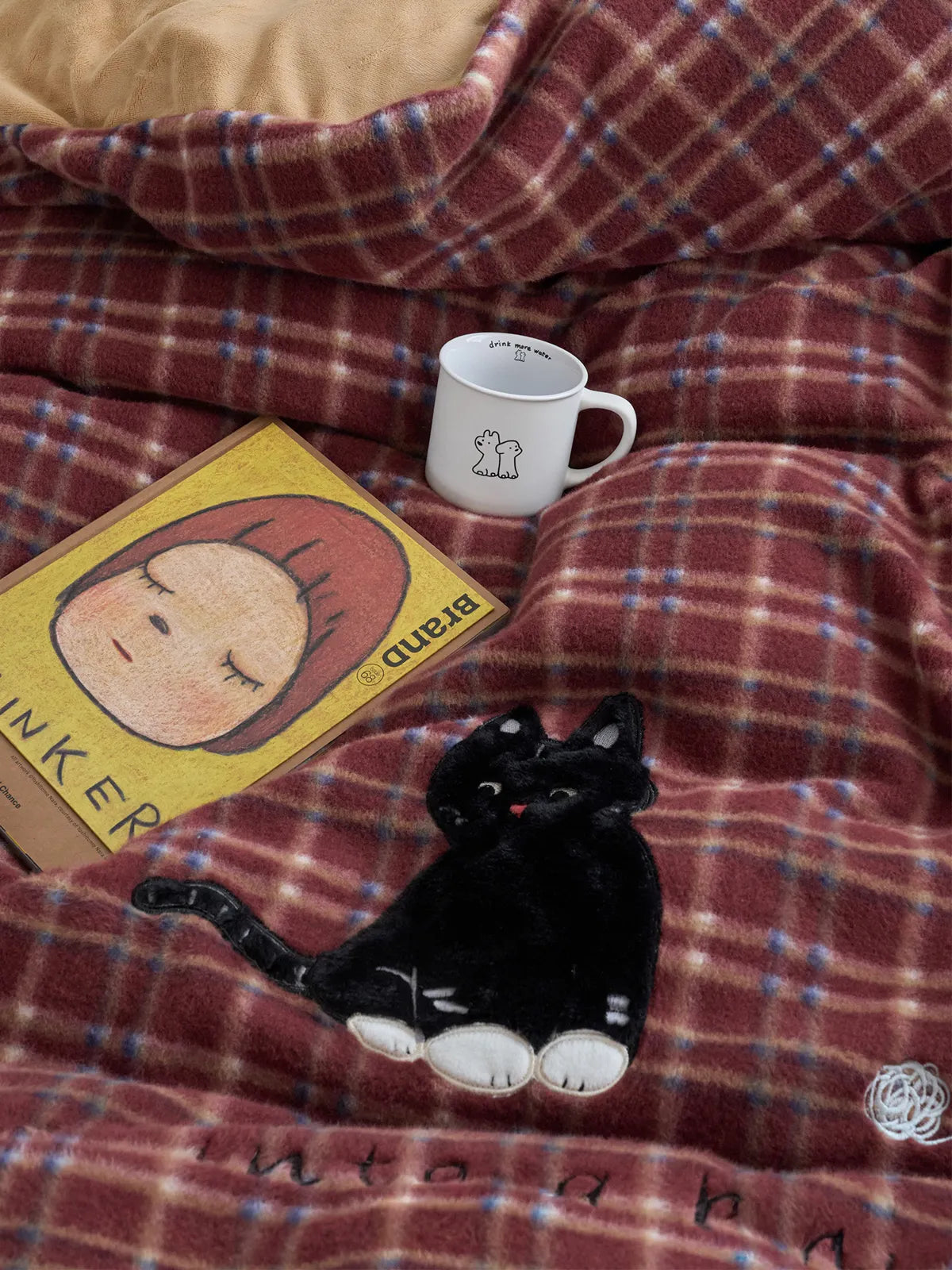 Overhead view of the red plaid bedding, showcasing the black cat embroidery, a cute mug, and a book. - RoomDen