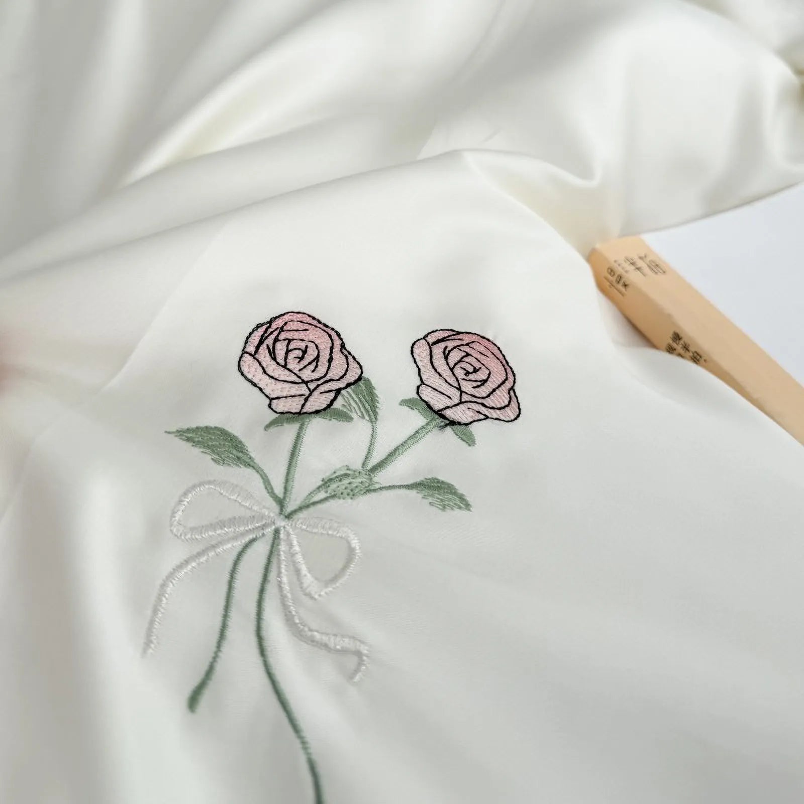 Macro shot of the embroidered two pink roses tied with a bow on the Cream White silky Tencel fabric. - RoomDen