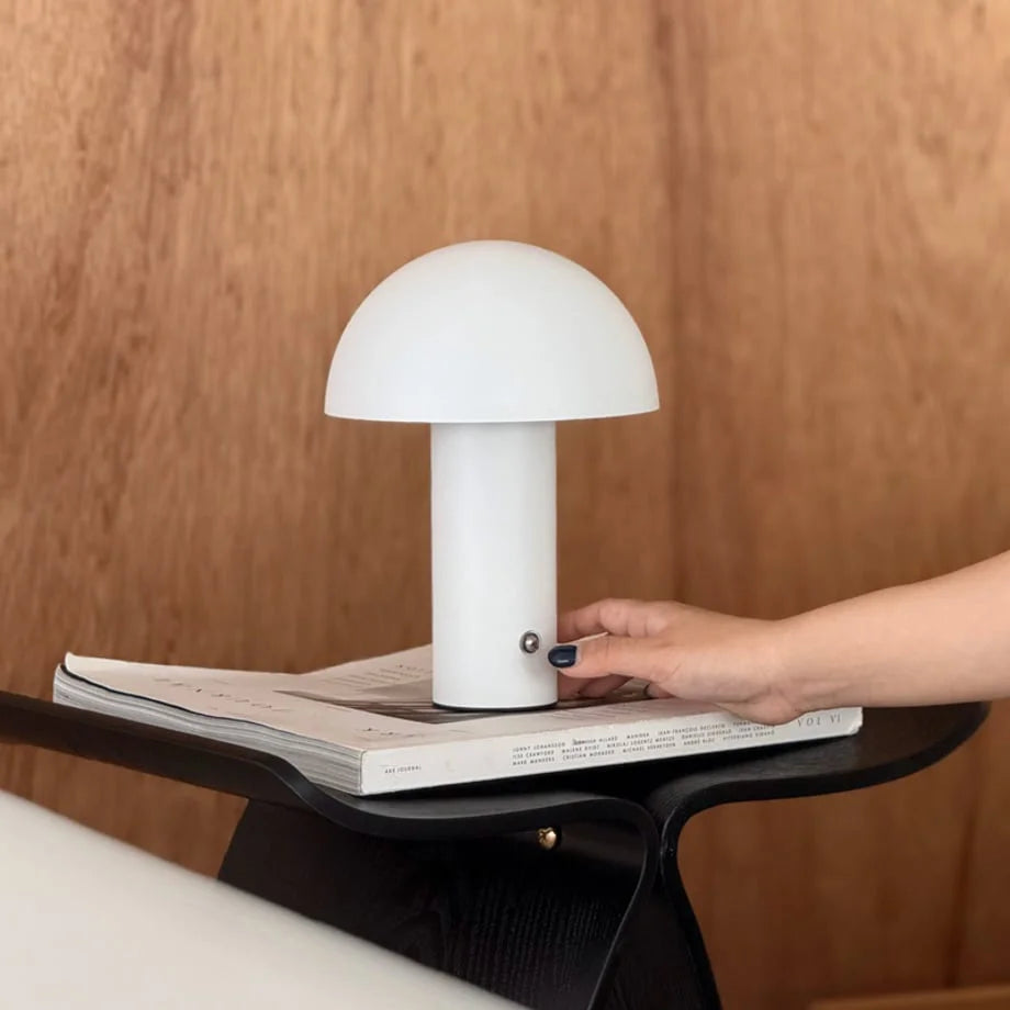 A hand reaching to touch the white version of the mushroom table lamp, resting on a magazine - RoomDen.