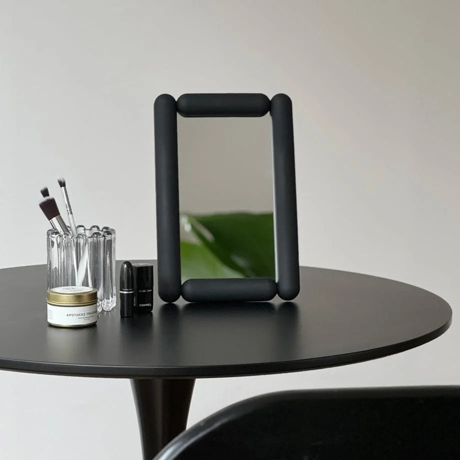 Black pill-shaped desktop mirror on a round table with makeup brushes. - RoomDen