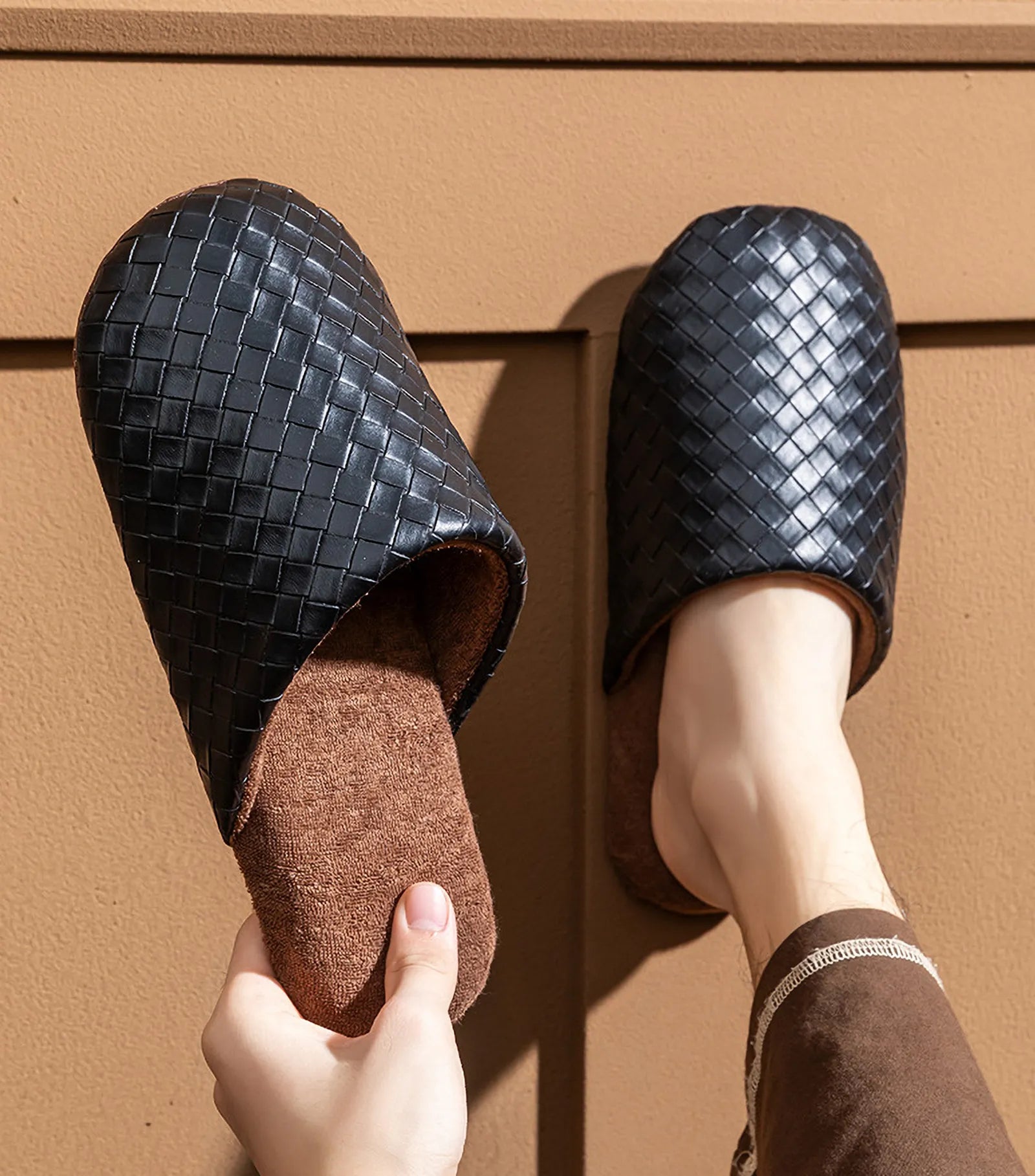 Model holding a black woven slipper against a brown wall background - RoomDen
