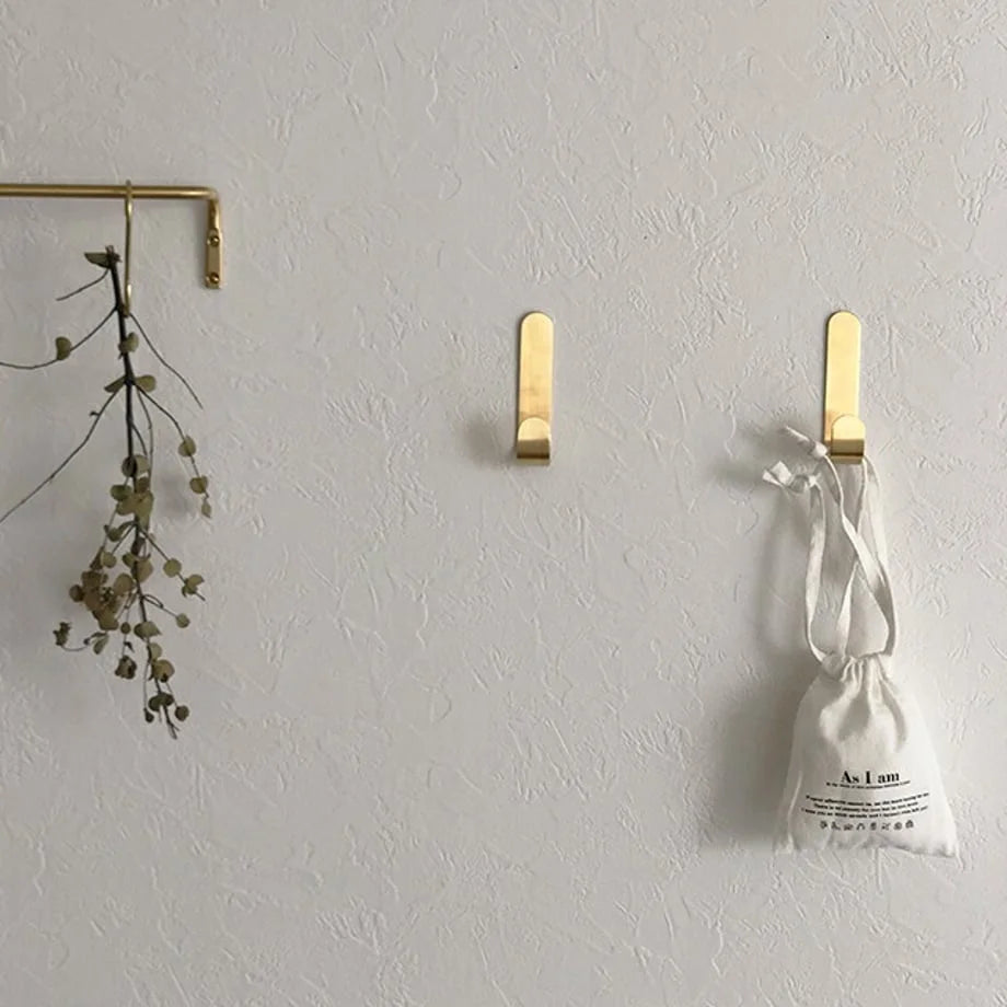 Lifestyle view of three minimalist brushed gold adhesive wall hooks on a white wall, holding a dried plant and a small bag - RoomDen