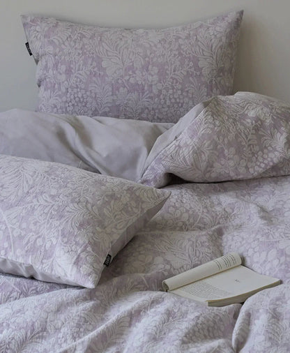 The dusty lavender Jacquard Botanical Bedding Set styled on a bed with an open book, showcasing the pillowcases - RoomDen.