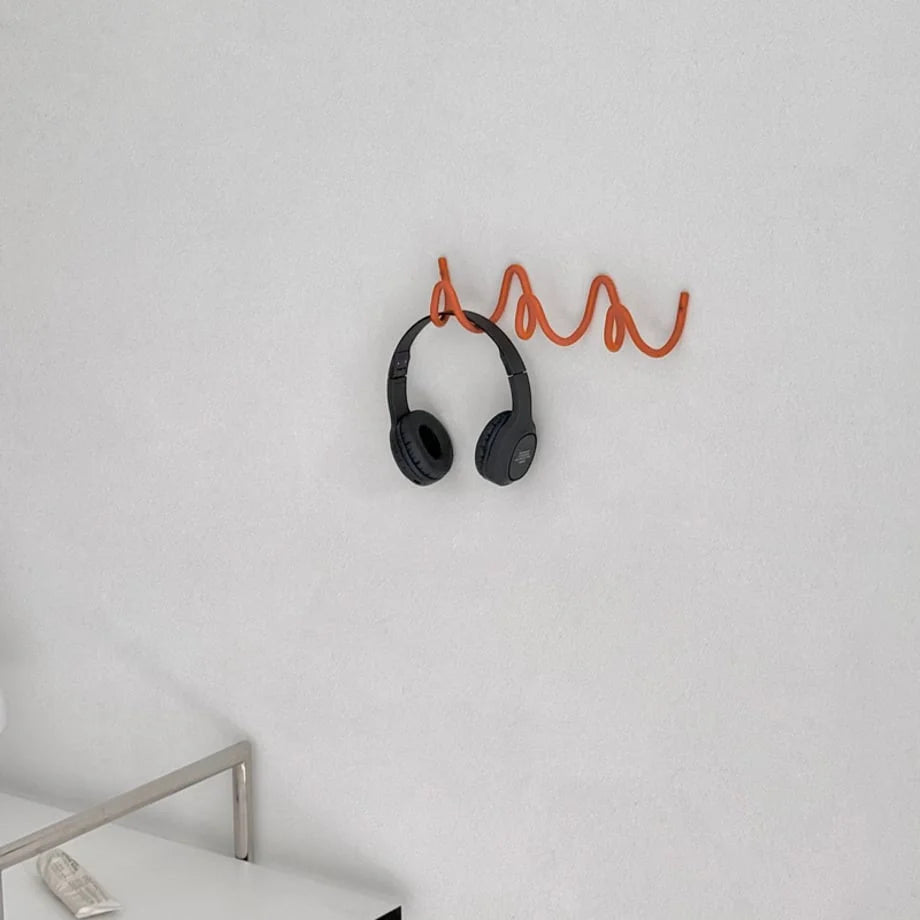 Angled view of black headphones on the orange Wavy Coil Metal Wall Hook against a white wall. - RoomDen
