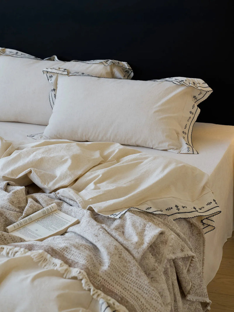 A cozy bedroom scene with the embroidered ruffle linen bedding styled with an open book and a knitted throw blanket. — RoomDen