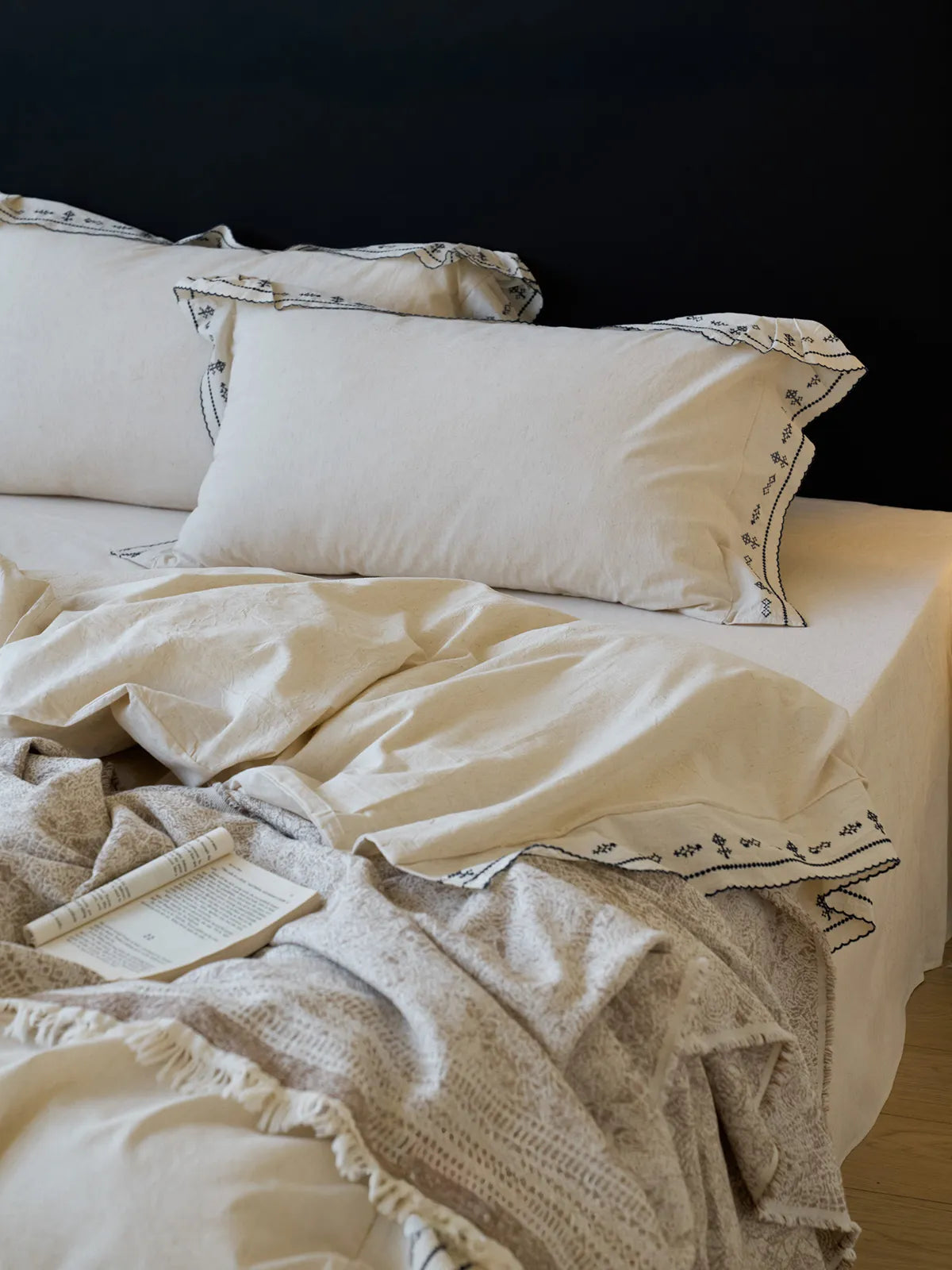 A cozy bedroom scene with the Embroidered Ruffle Linen Bedding Set, styled with an open book and a knitted throw blanket - RoomDen.