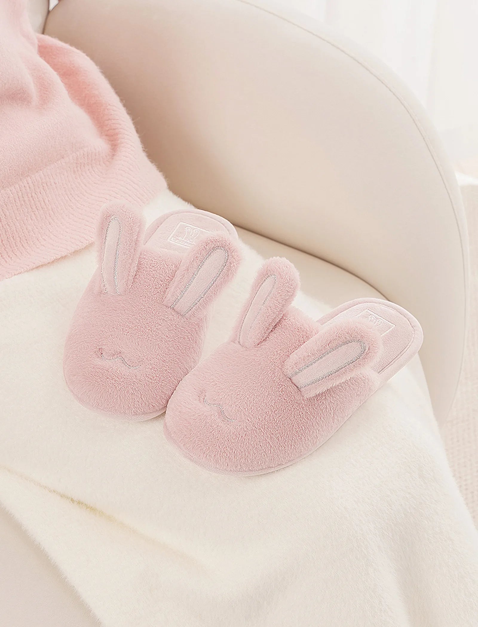 Pair of pink faux fur bunny slippers on white chair with sweater - RoomDen