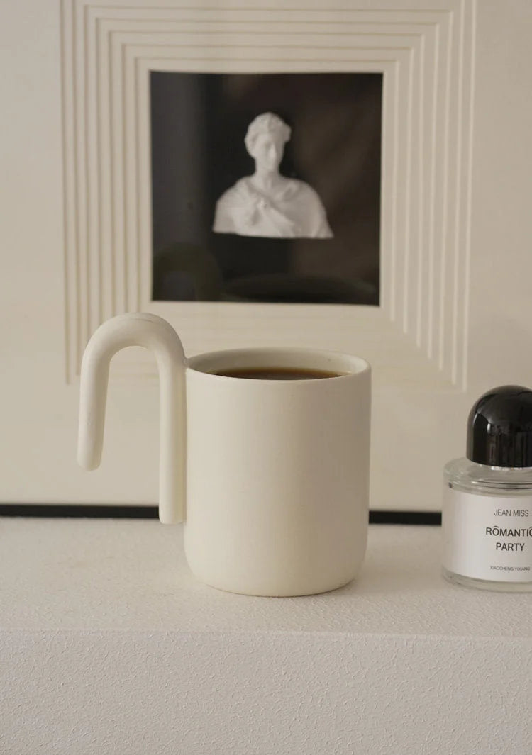 Cream colored minimalist mug with sculptural handle on a white mantelpiece - RoomDen
