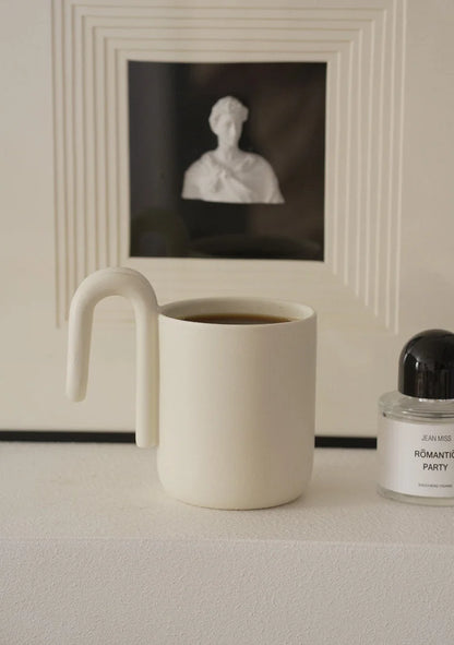 Cream colored minimalist mug with sculptural handle on a white mantelpiece - RoomDen