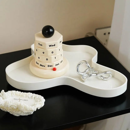 The wooden perpetual calendar placed on a modern, wavy white tray next to a silver sculpture. - RoomDen