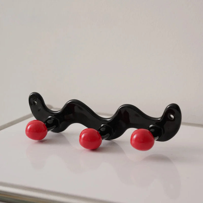 Three-hook wavy black ceramic wall rack with red balls on a white surface.