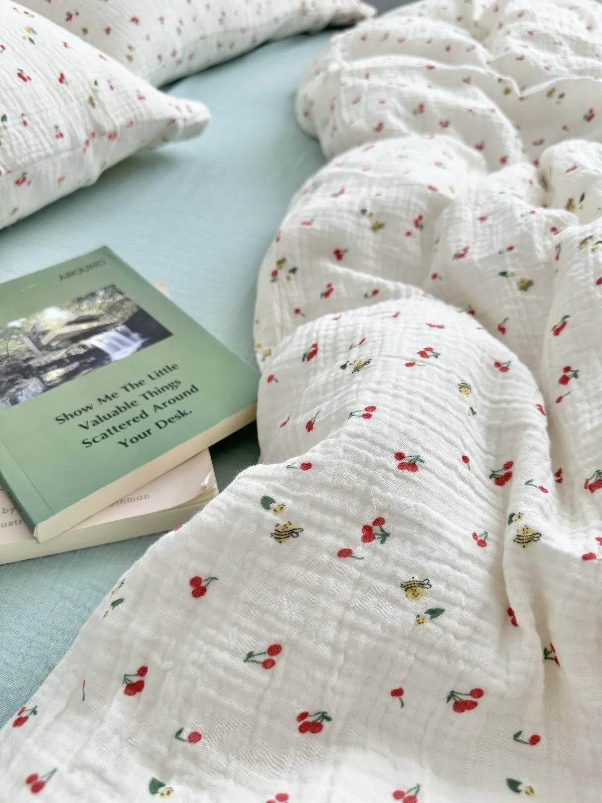 Close up of cream textured duvet cover with tiny cherry and bee print, styled with a green book titled "Show Me The Little Valuable Things". RoomDen.
