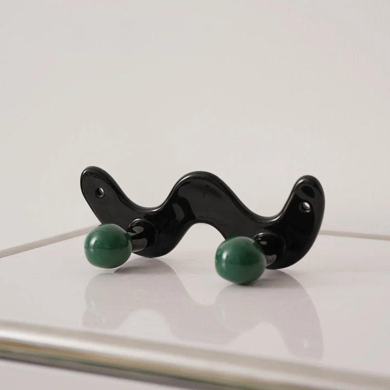 Glossy black wavy wall hook with two green ceramic balls on a white surface - RoomDen.