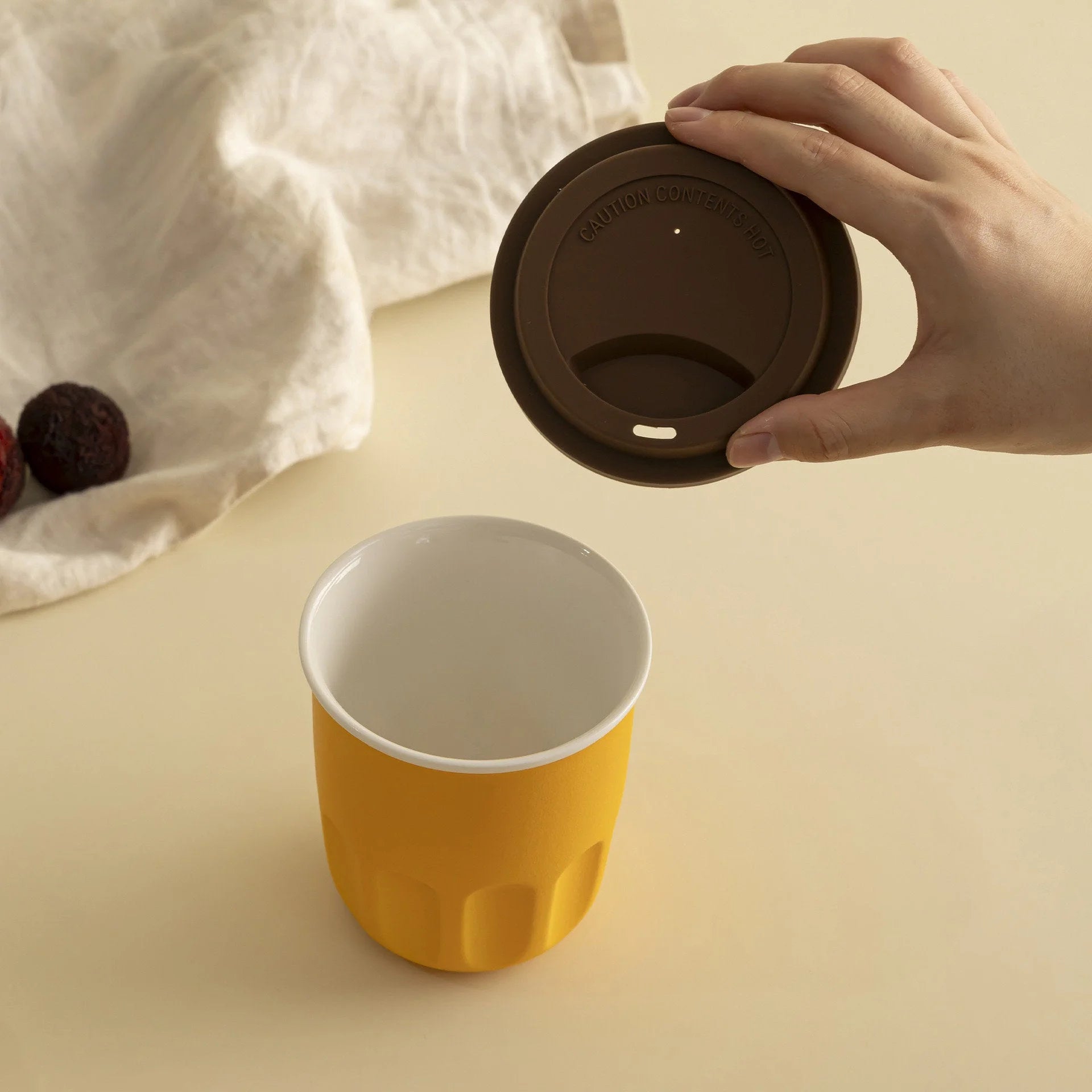 Hand placing a brown silicone lid on a mustard yellow frosted ceramic reusable coffee cup. - RoomDen