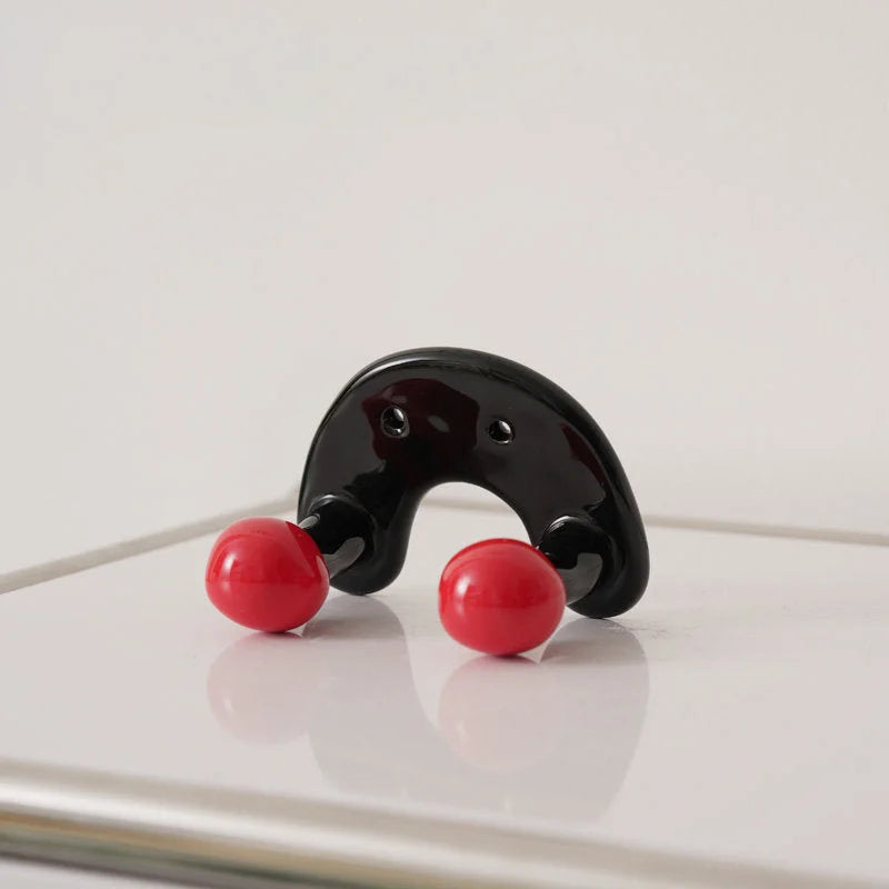 U-shaped black ceramic wall hook with two red balls on a reflective white surface.