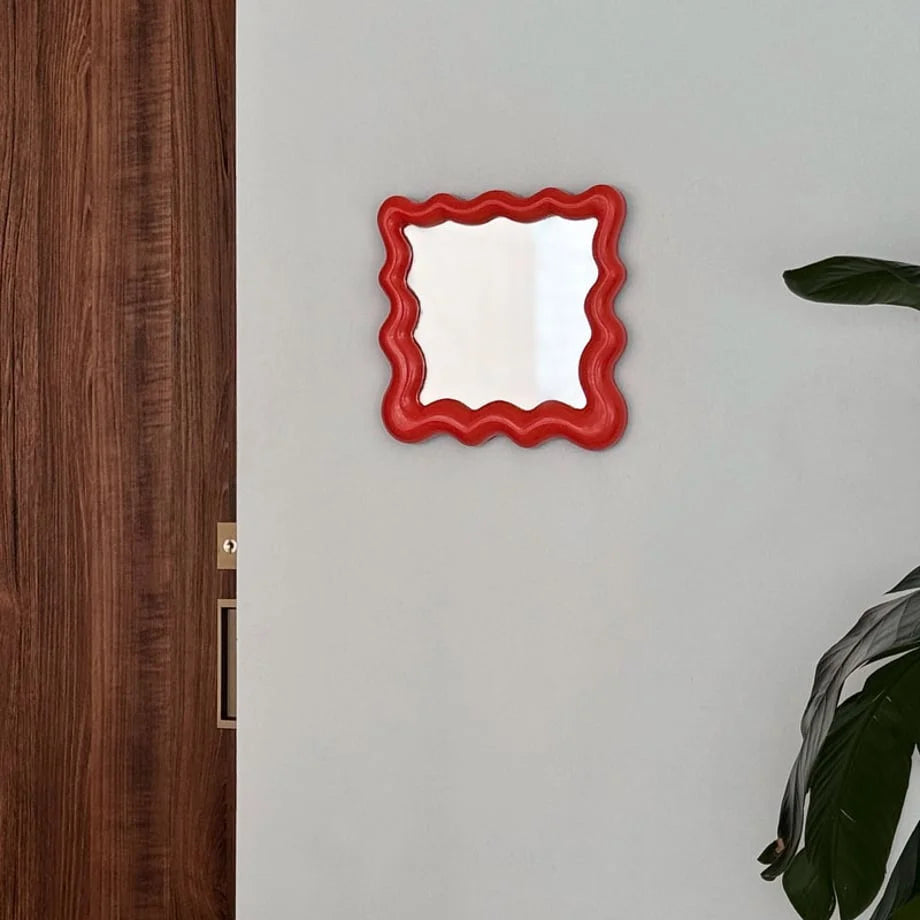 Vibrant red wavy mirror hanging on a wall next to a wooden door and a houseplant - RoomDen