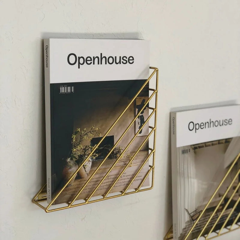 Slim wireframe metal magazine rack holding a magazine against a pale reading-corner wall - RoomDen