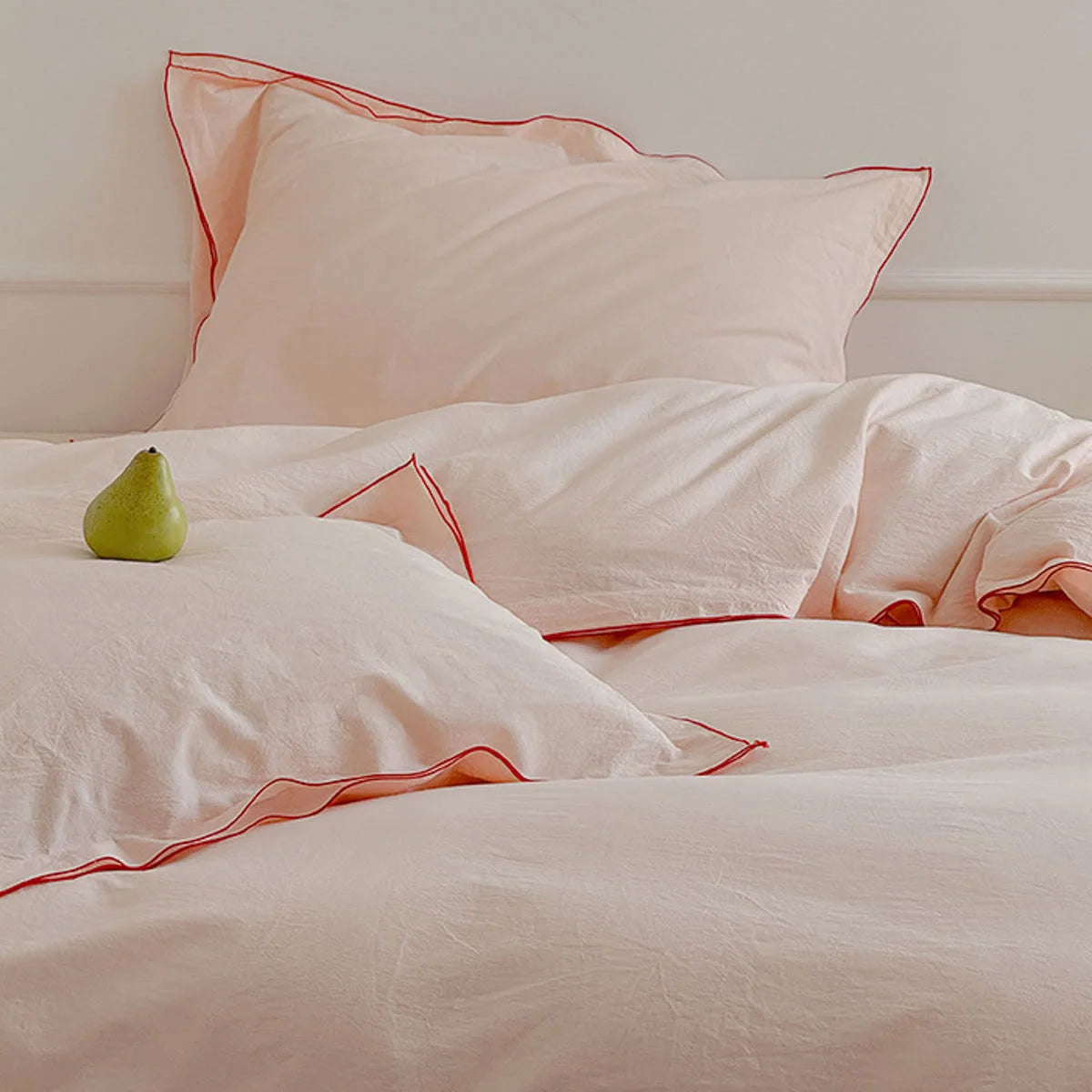 Blush pink washed cotton bedding set with red ruffle trim, with a single green pear placed on the pillow.