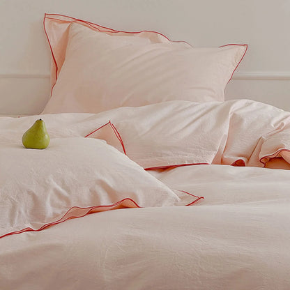 Blush pink washed cotton bedding set with red ruffle trim, with a single green pear placed on the pillow.