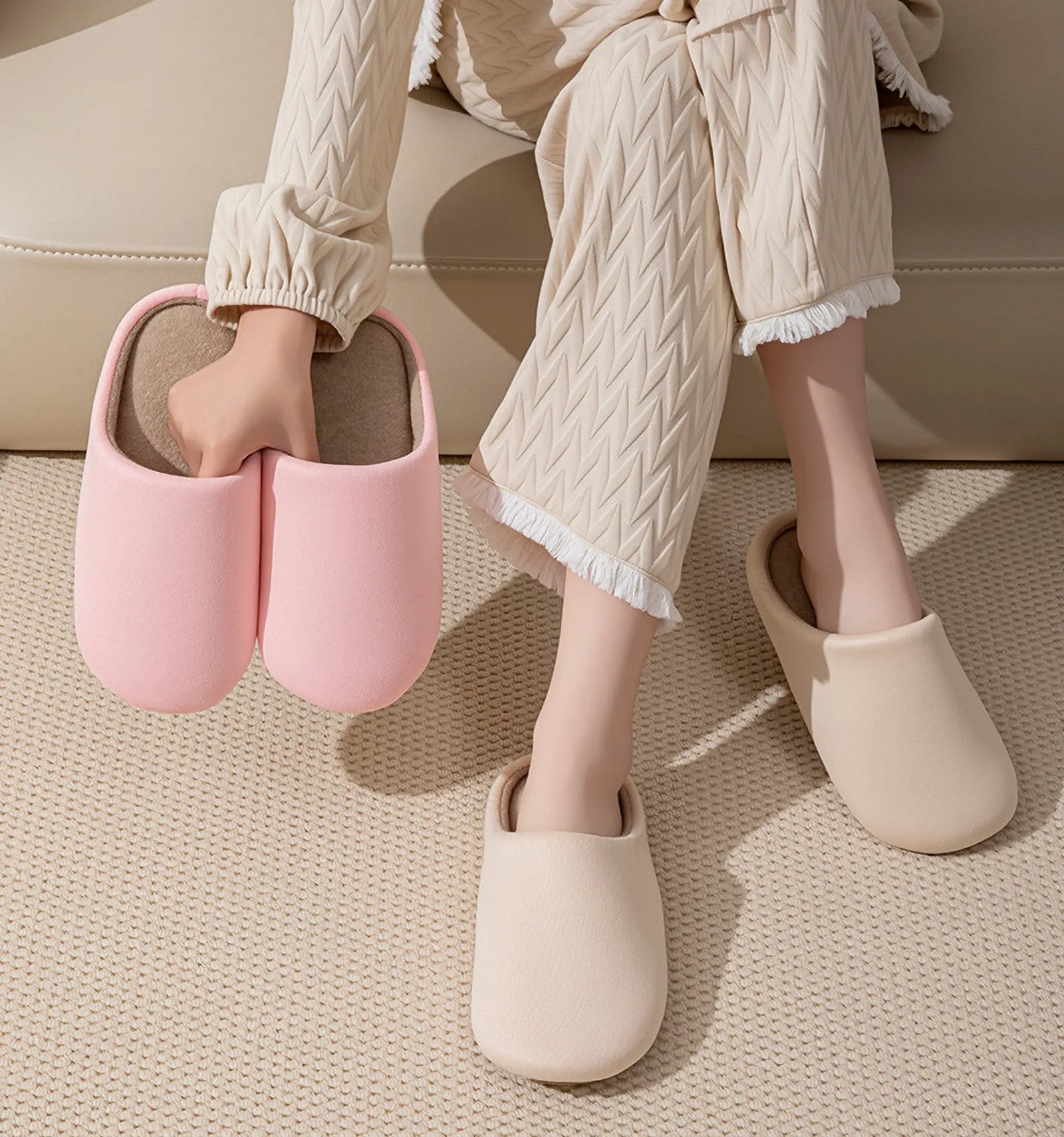 Hands holding pink slippers showing soft texture with beige pair on feet - RoomDen