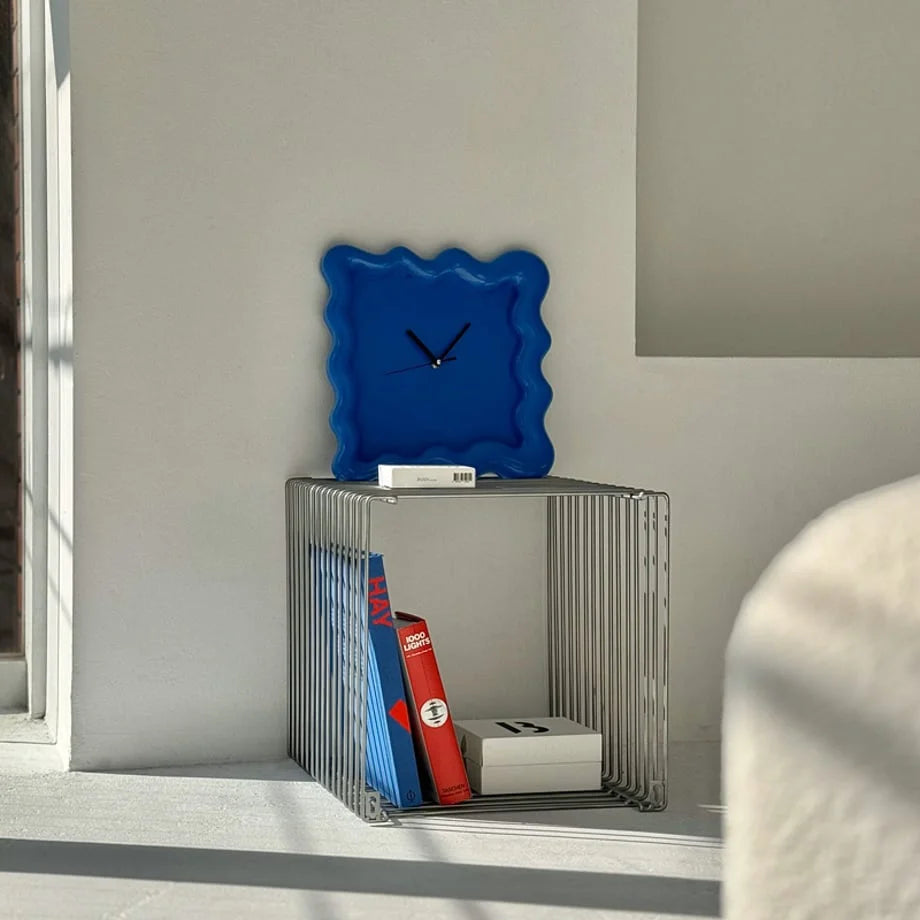 A royal blue Wavy Squiggle Clock placed on a wireframe metal side table next to books. - RoomDen