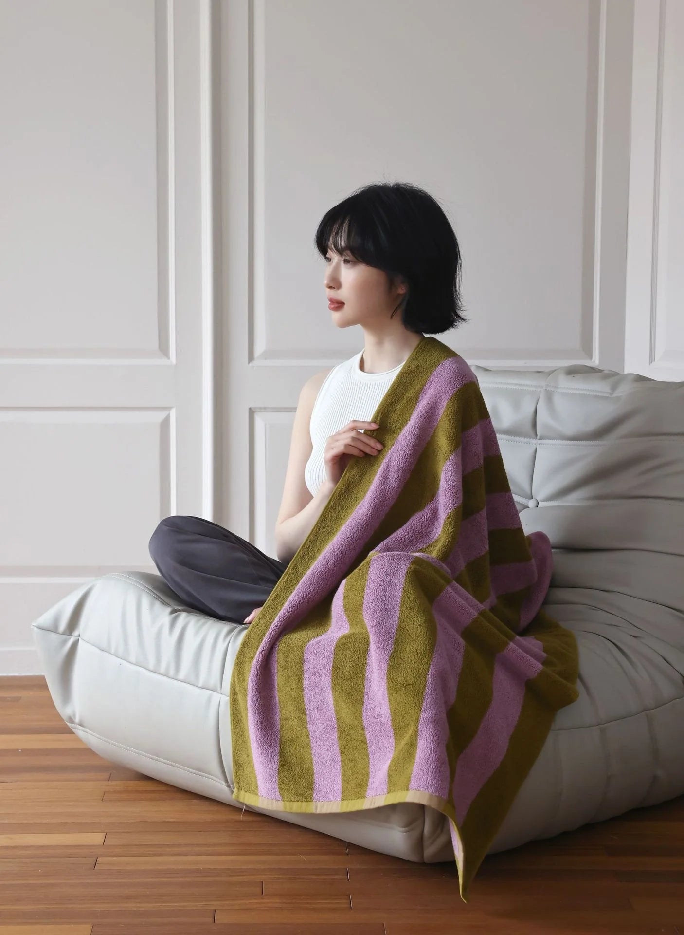 A woman wrapped in the green and purple Bold Striped Cotton Bath Towel, sitting on a modern grey lounge chair. - RoomDen