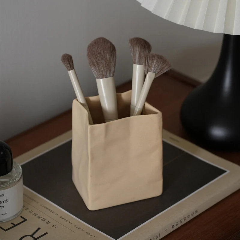 A khaki crumpled ceramic organizer holding makeup brushes, sitting on a book on a wooden desk. - RoomDen