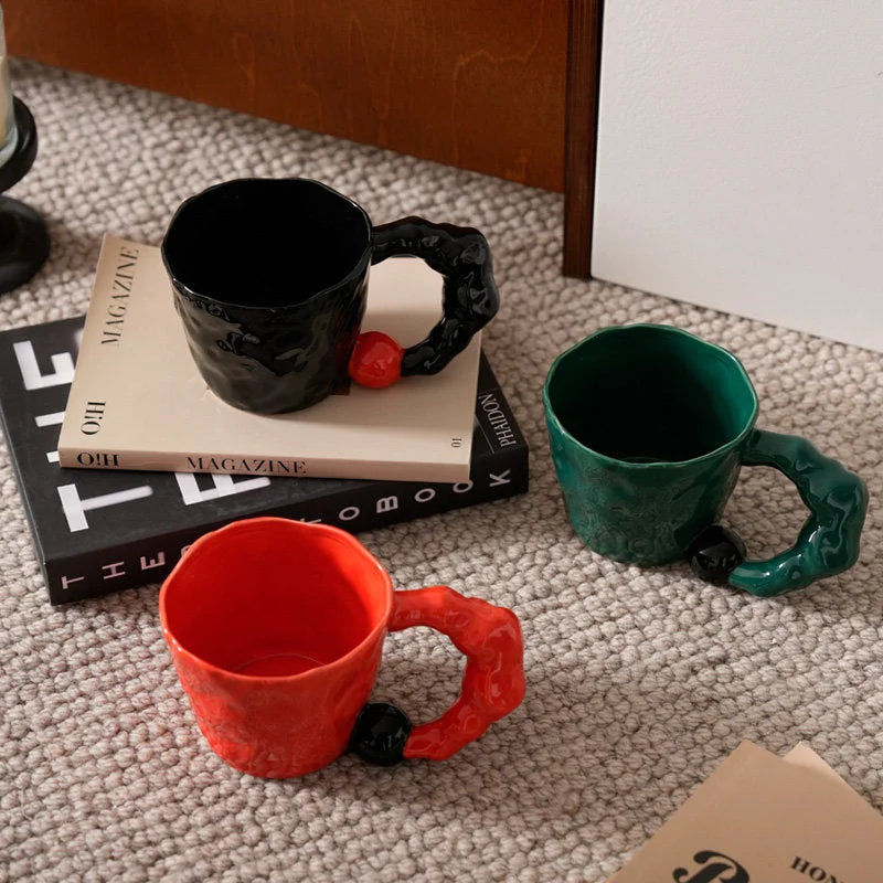 Top-down view of black, red, and green chunky ceramic mugs arranged on a textured rug with magazines - RoomDen.