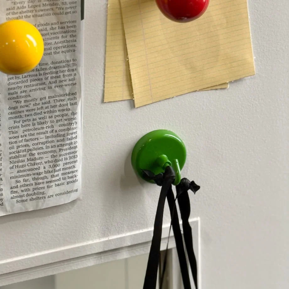 Green magnetic hook holding black straps on a white refrigerator door next to post-it notes - RoomDen