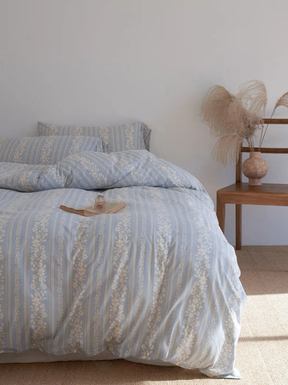 The cornflower blue floral stripe cotton bedding set on a bed next to a wooden side table with dried pampas grass. - RoomDen