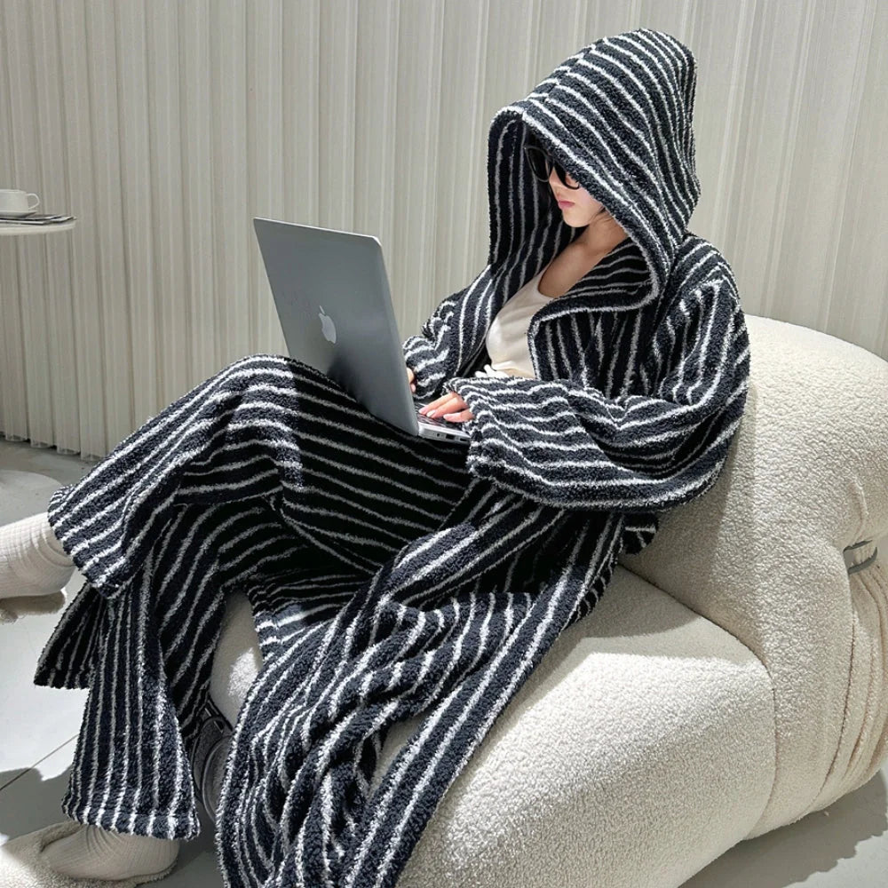 Model sitting on a white bouclé chair, wearing a plush black and white striped hooded pajama set while working on a laptop - RoomDen.